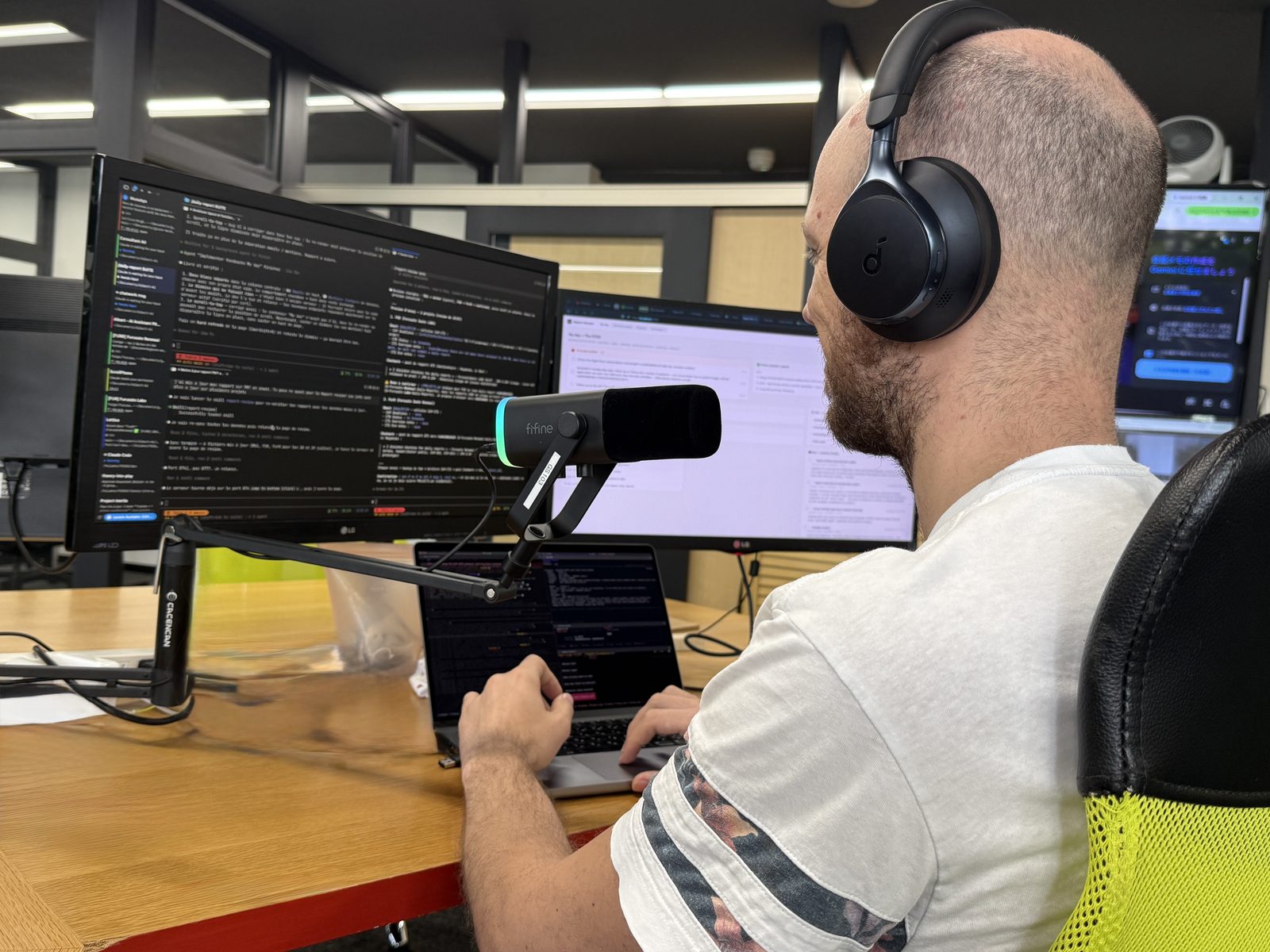 指向性マイクをアームで口元に寄せ、ヘッドホンをつけてノートパソコンに向かうエンジニア。隣にはコードとチャットを映した大型モニターが並んでいる